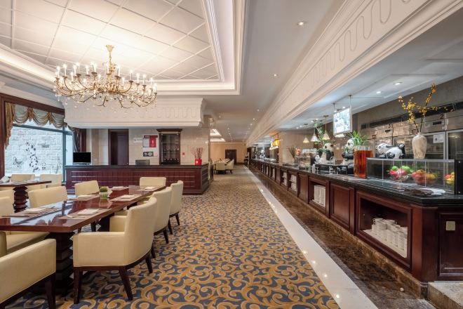 Um saguão de hotel com um balcão de recepção comprido, uma área de refeições com mesas e cadeiras, e um bar com exposição de frutas e flores.