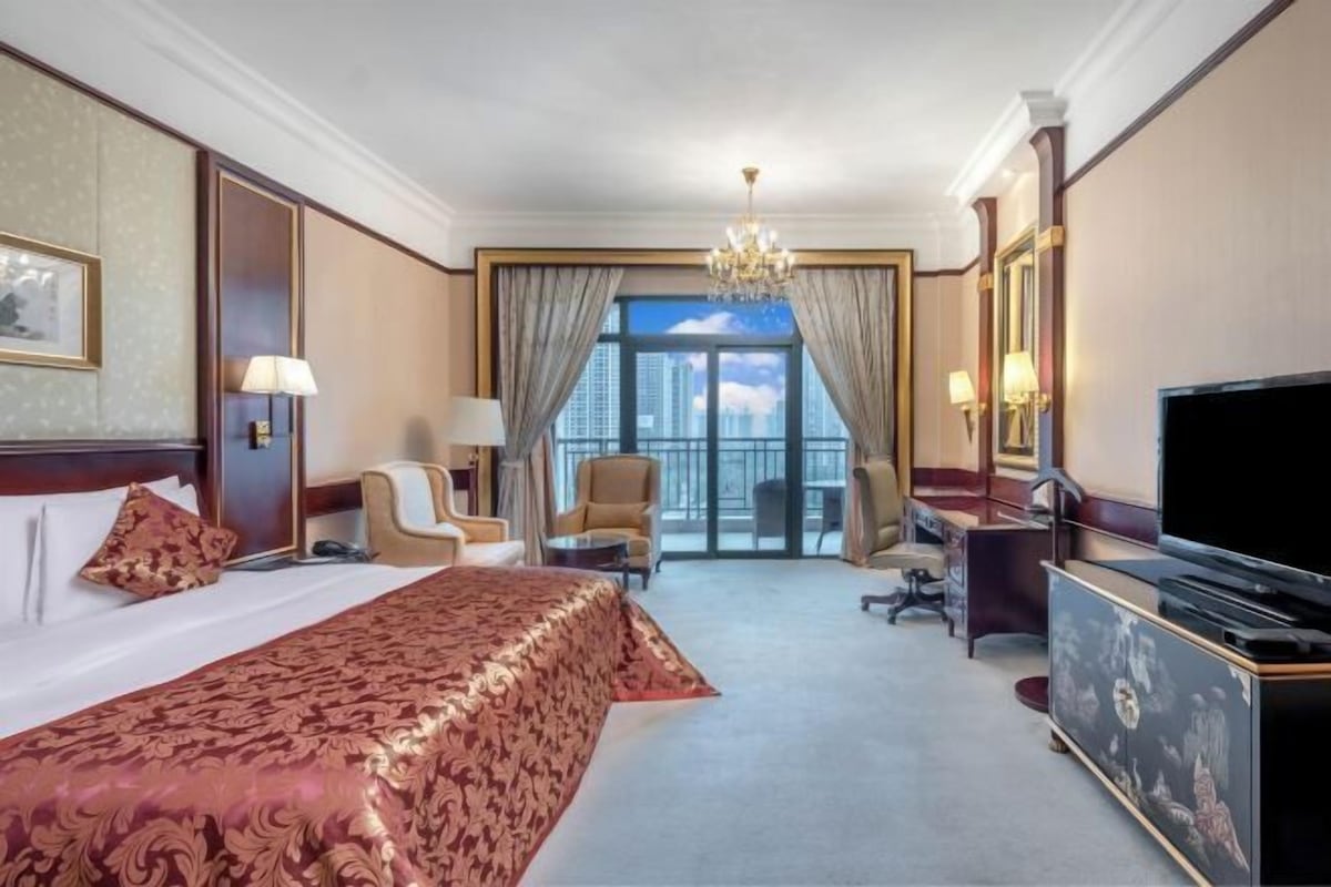A hotel room with a large bed, a TV, a desk, and a view of the city through the window.