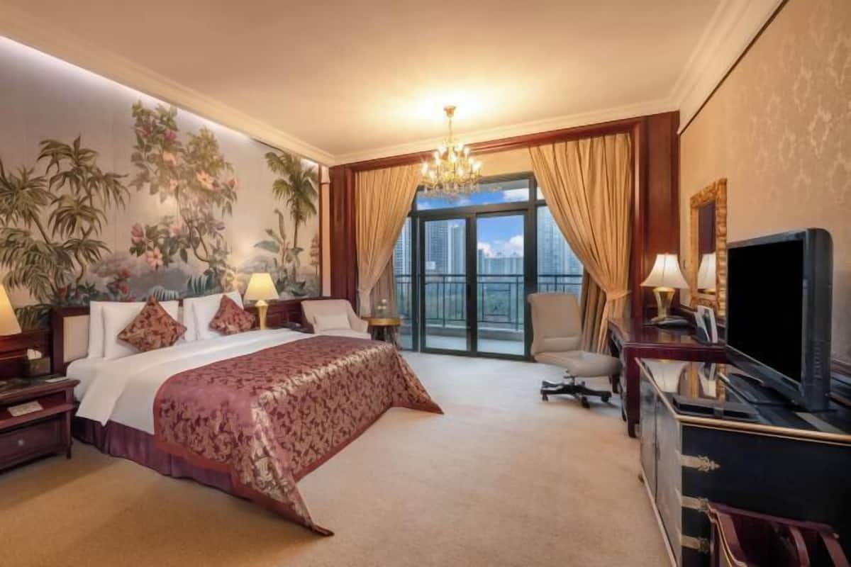 A hotel room with a large bed, a television, a desk, and a view of the city through the window.