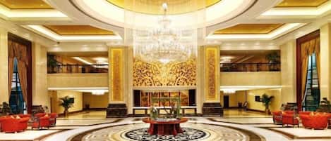 Um amplo hall com um lustre central, piso com padrão circular e área de estar com sofás vermelhos.