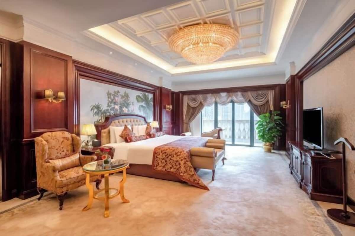 A spacious hotel room with a large bed, a sitting area with a chair and table, a television, and a chandelier.