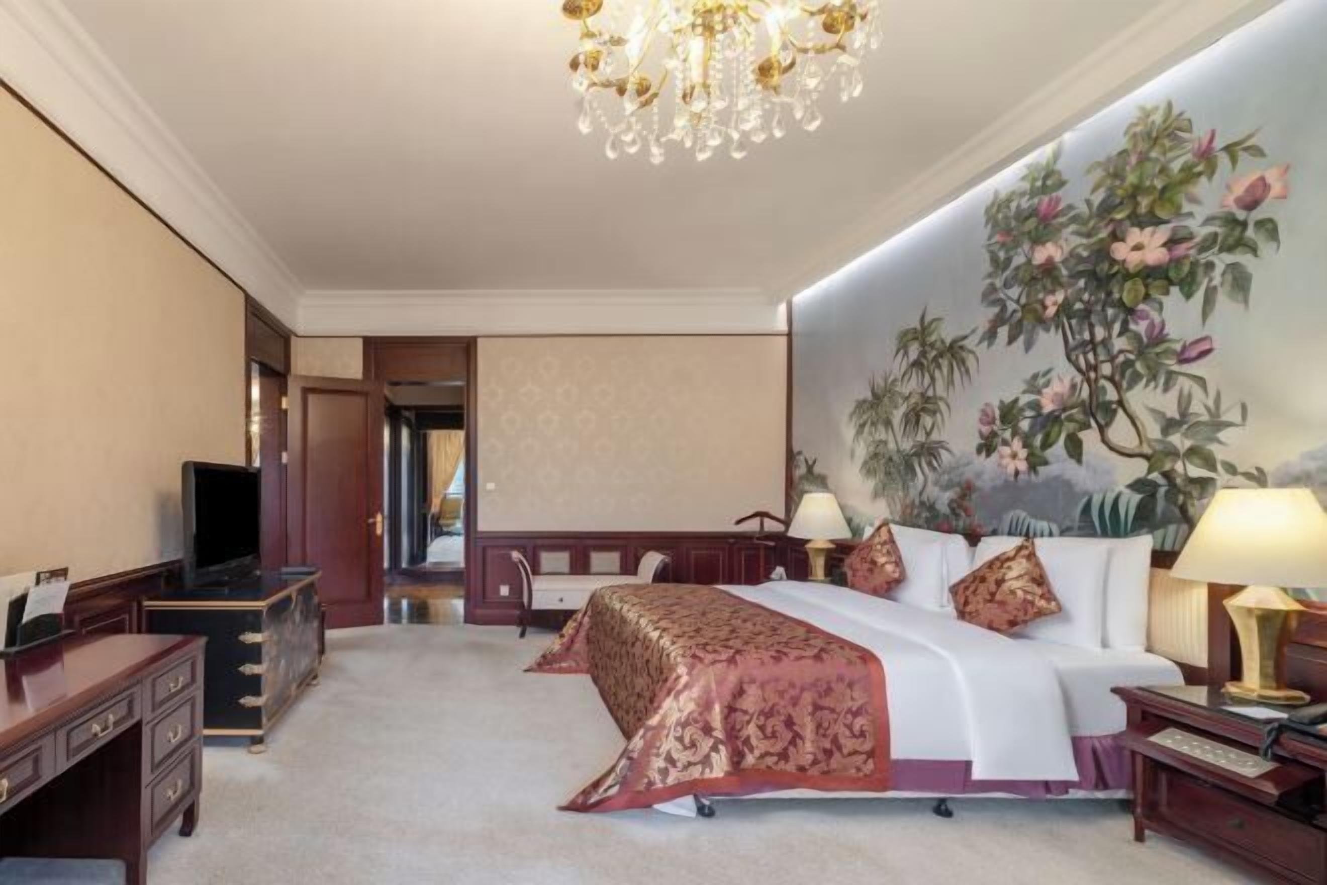 A hotel room with a large bed, a desk, a television, and a chandelier.