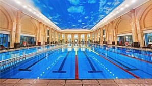 Una piscina interior con un mural en el techo que representa un cielo azul y nubes, rodeada de columnas en arco y grandes ventanales.