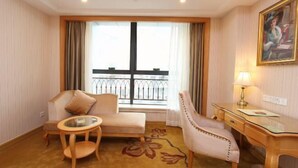 A hotel room with a sofa, armchair, small round table, and a desk with a lamp. There is a large window with a view of the city. - Vienna International Hotel (Shenzhen Xiashuijing Metro Station) (Shenzhen)
