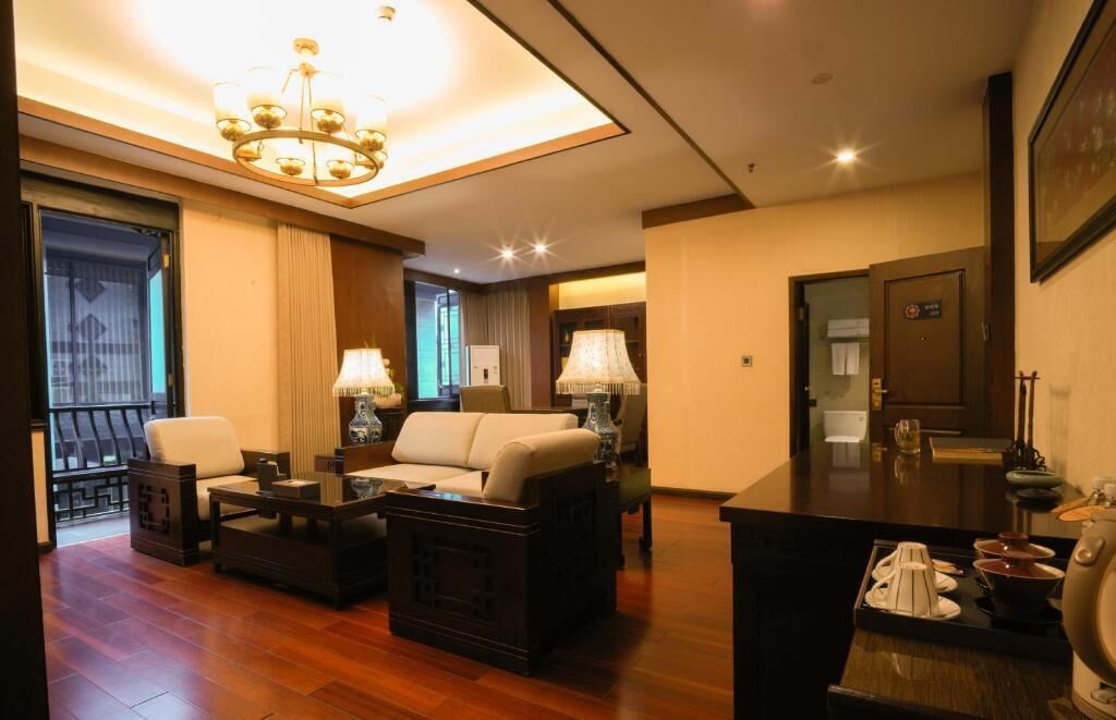 Habitación de hotel con sala de estar que incluye un sofá, dos sillones, una mesa de centro y una mesa de comedor con una lámpara. Hay un balcón con barandilla y una puerta a la derecha.