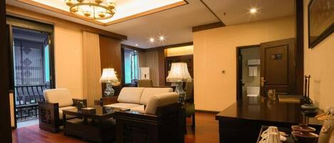 Habitación de hotel con sala de estar que incluye un sofá, dos sillones, una mesa de centro y una mesa de comedor con una lámpara. Hay un balcón con barandilla y una puerta a la derecha.