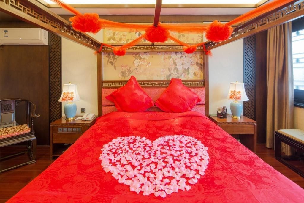 A hotel room with a red bedspread, a heart-shaped arrangement of rose petals, and traditional decor.