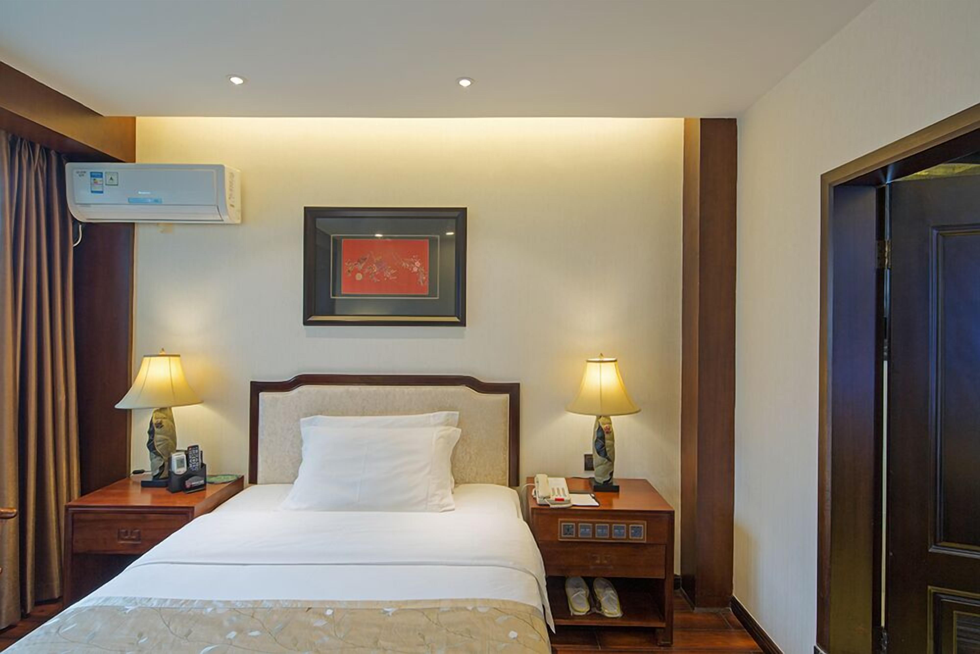 A hotel room with a bed, two bedside tables with lamps, a framed artwork above the bed, an air conditioning unit, and a door.