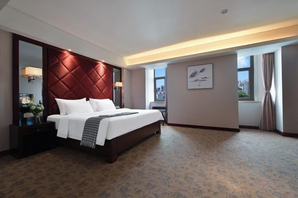 A hotel room with a large bed, a red headboard, a nightstand with a lamp, a window with curtains, and a framed picture on the wall.