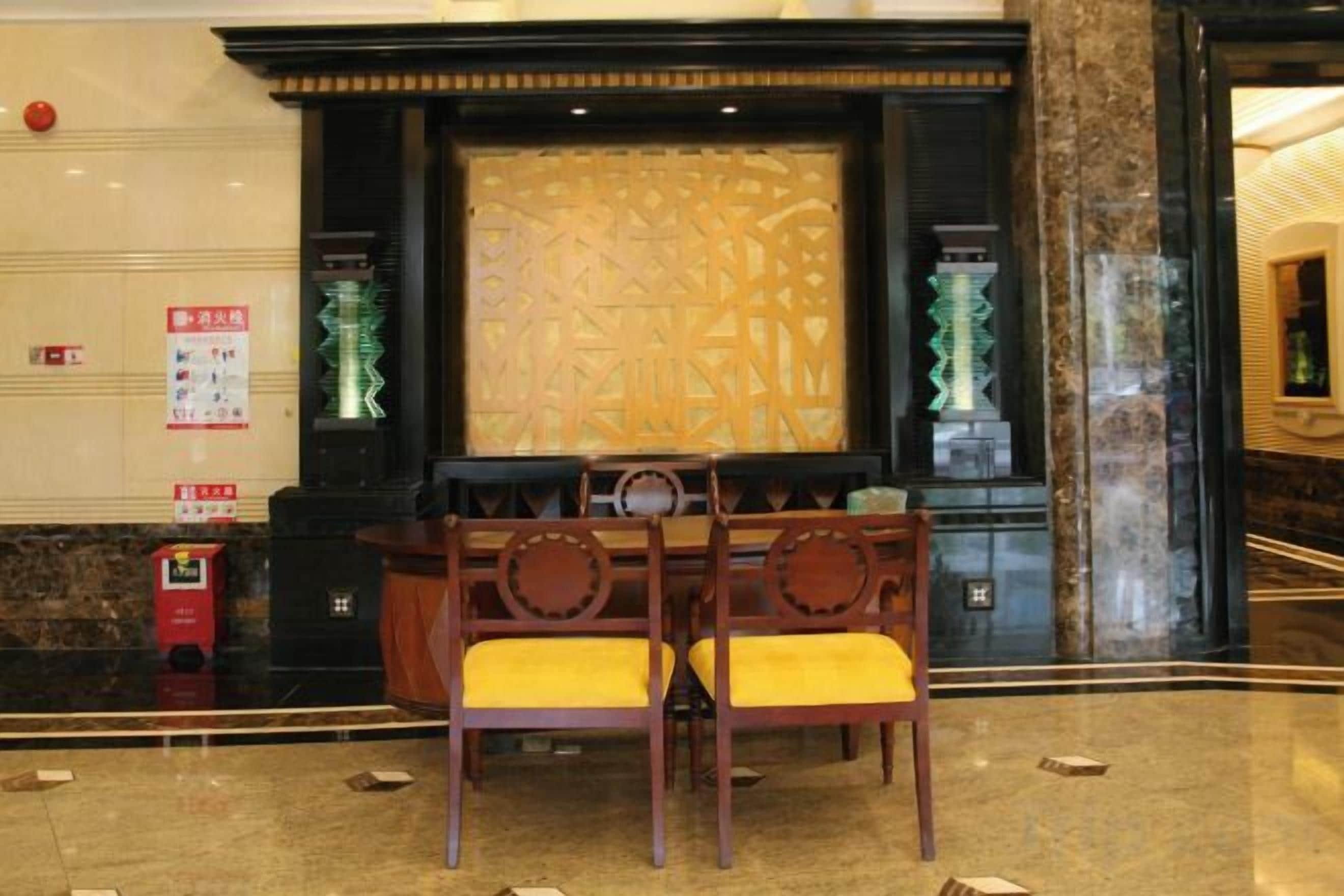 A lobby with a reception desk, two wooden chairs with yellow cushions, a decorative wall, and a fire extinguisher.