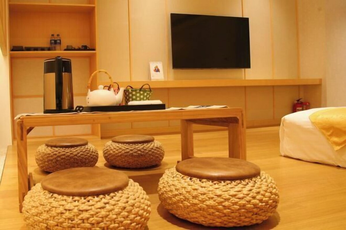A hotel room with a wooden table, four woven stools, a television, and a bed with a yellow blanket.
