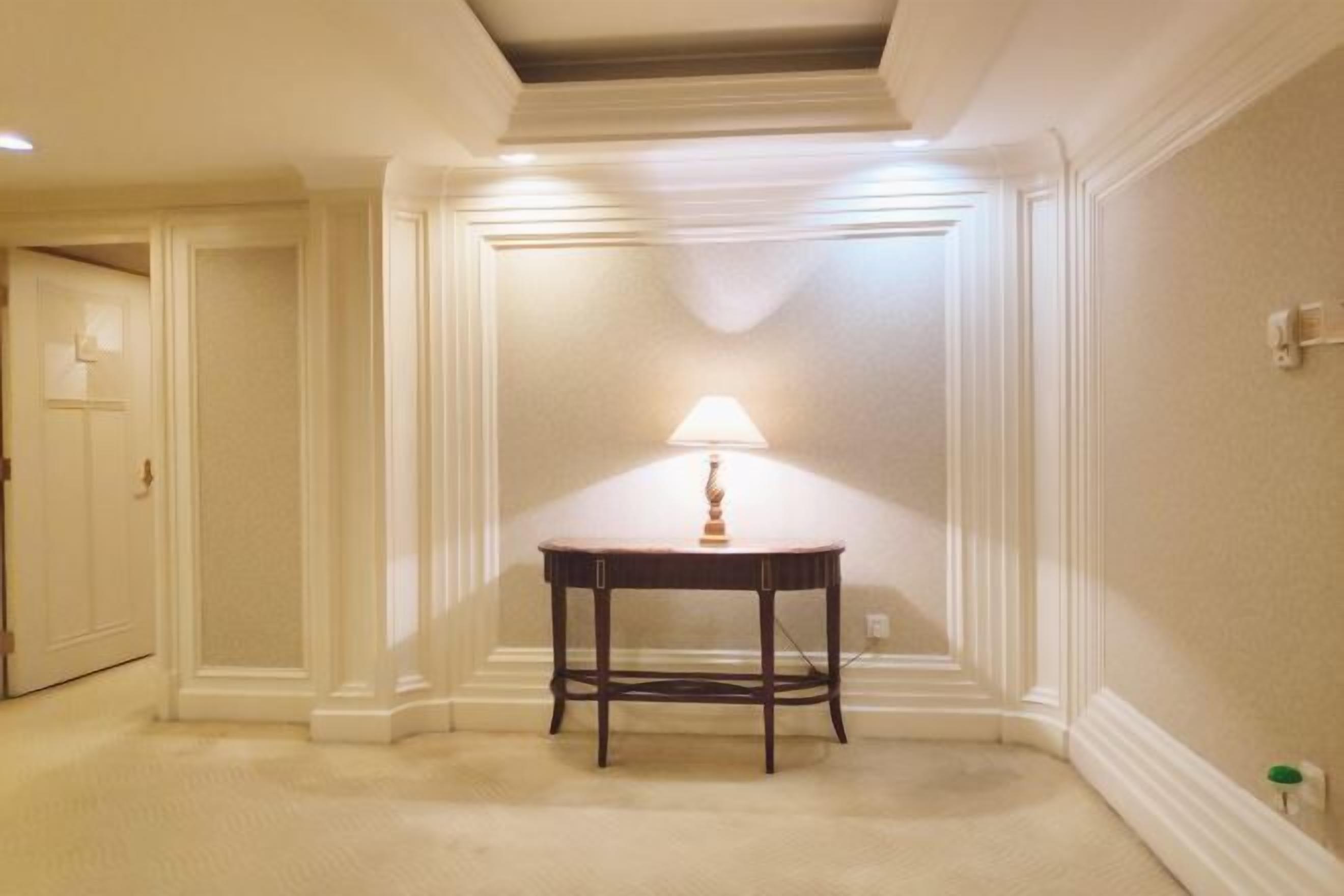 A hotel room with a carpeted floor, a table with a lamp, and a door in the background.