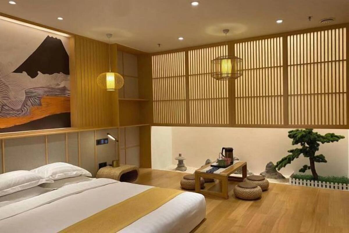 A hotel room with a bed, a desk, a lamp, a wall-mounted shelf, and a decorative wall piece.