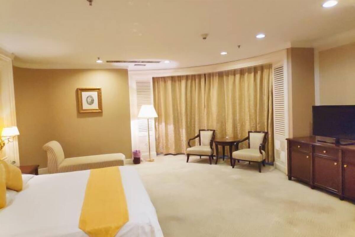 A hotel room with a large bed, two armchairs, a small table, a TV, and a window with curtains.