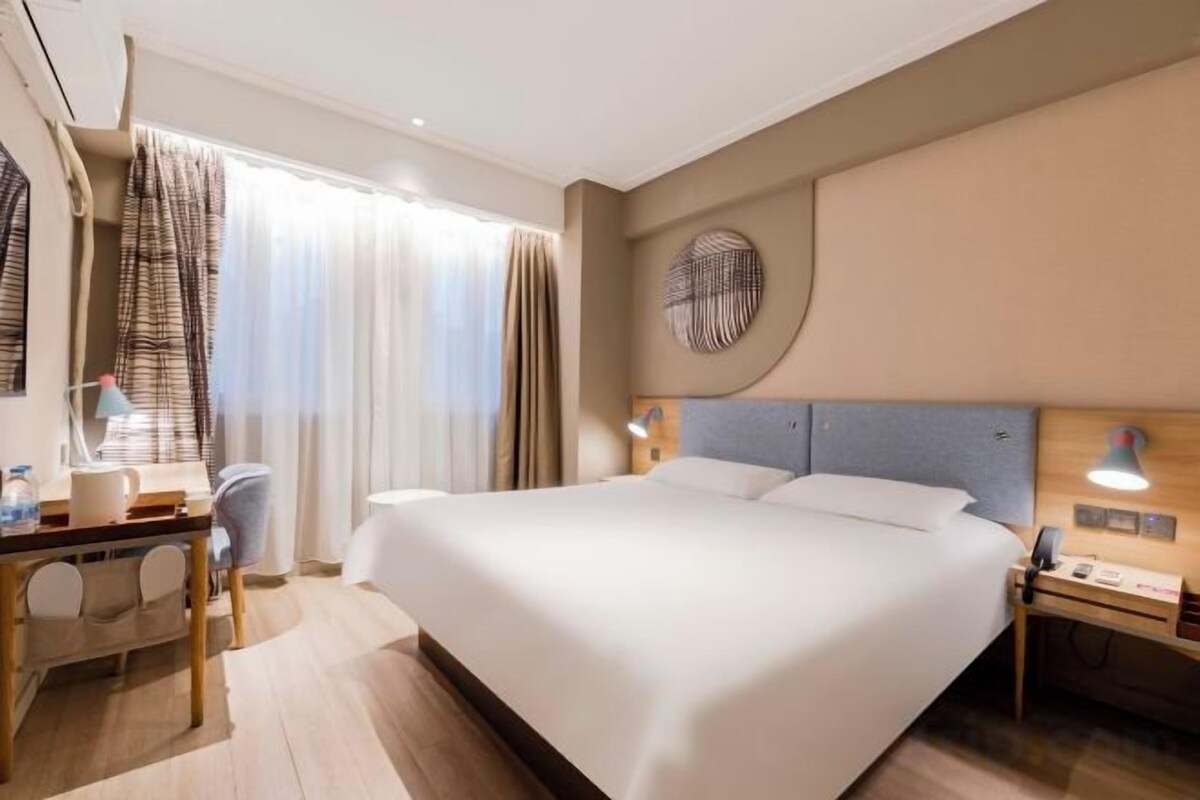 A hotel room with a large bed, bedside tables, a desk with a chair, and a window with curtains.
