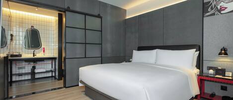 A modern hotel room with a large bed, a built-in desk, a vanity with a mirror, and a red nightstand.