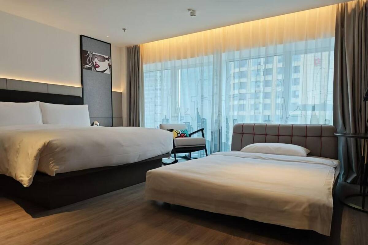 A hotel room with two beds, a sofa, a chair, and a large window with a city view.
