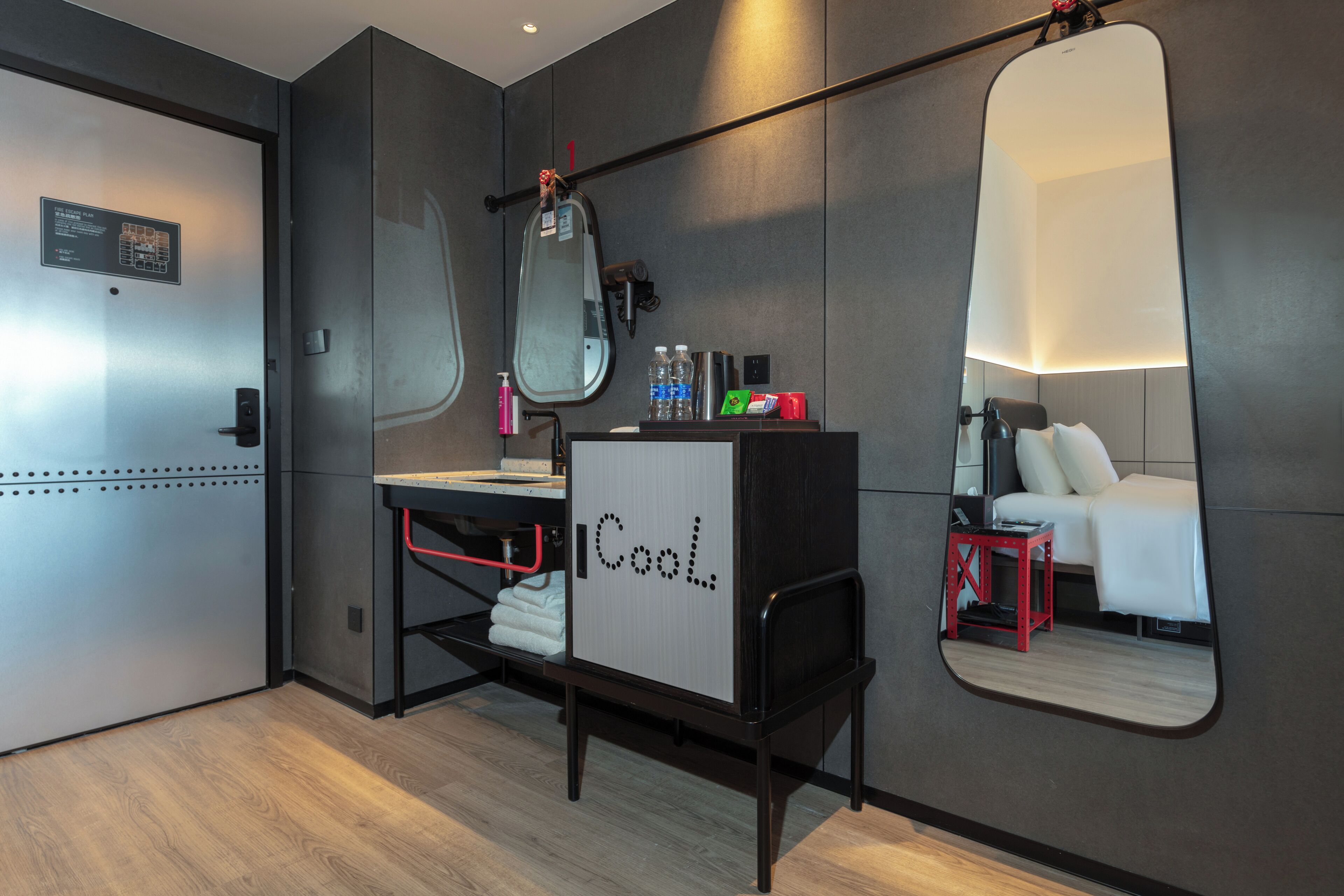 Una habitación de hotel moderna con escritorio, espejo, cama y un taburete pequeño rojo.