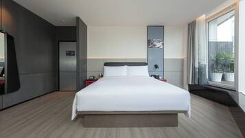Quarto de hotel moderno com uma cama grande, um guarda-roupa embutido, uma pequena mesa de cabeceira e uma janela ampla com vista para o exterior.