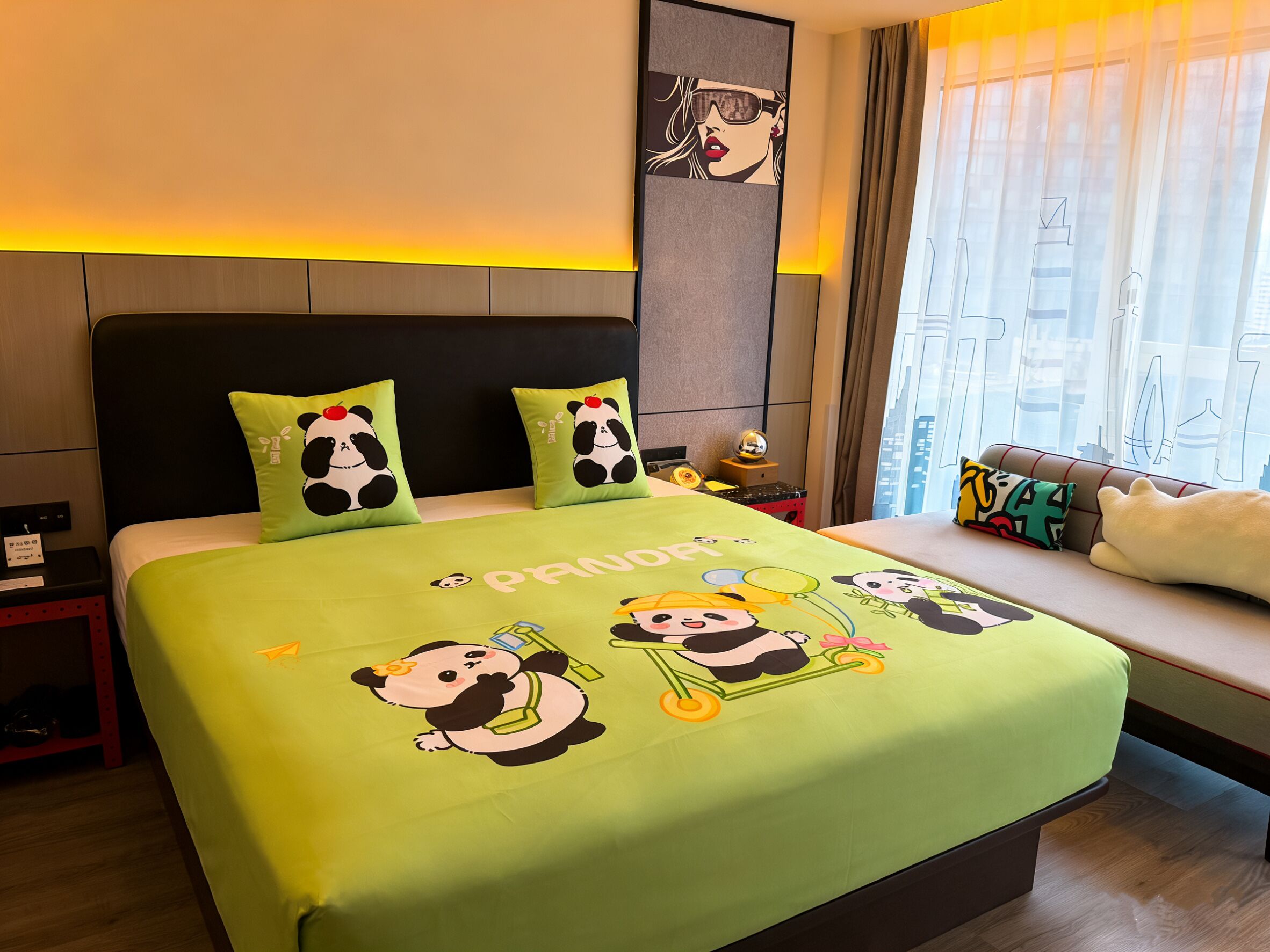 Quarto de hotel com uma cama e roupa de cama com tema de panda, mesa de cabeceira com luminária e janela com cortinas.