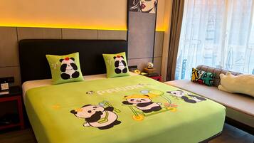 Quarto de hotel com uma cama e roupa de cama com tema de panda, mesa de cabeceira com luminária e janela com cortinas.