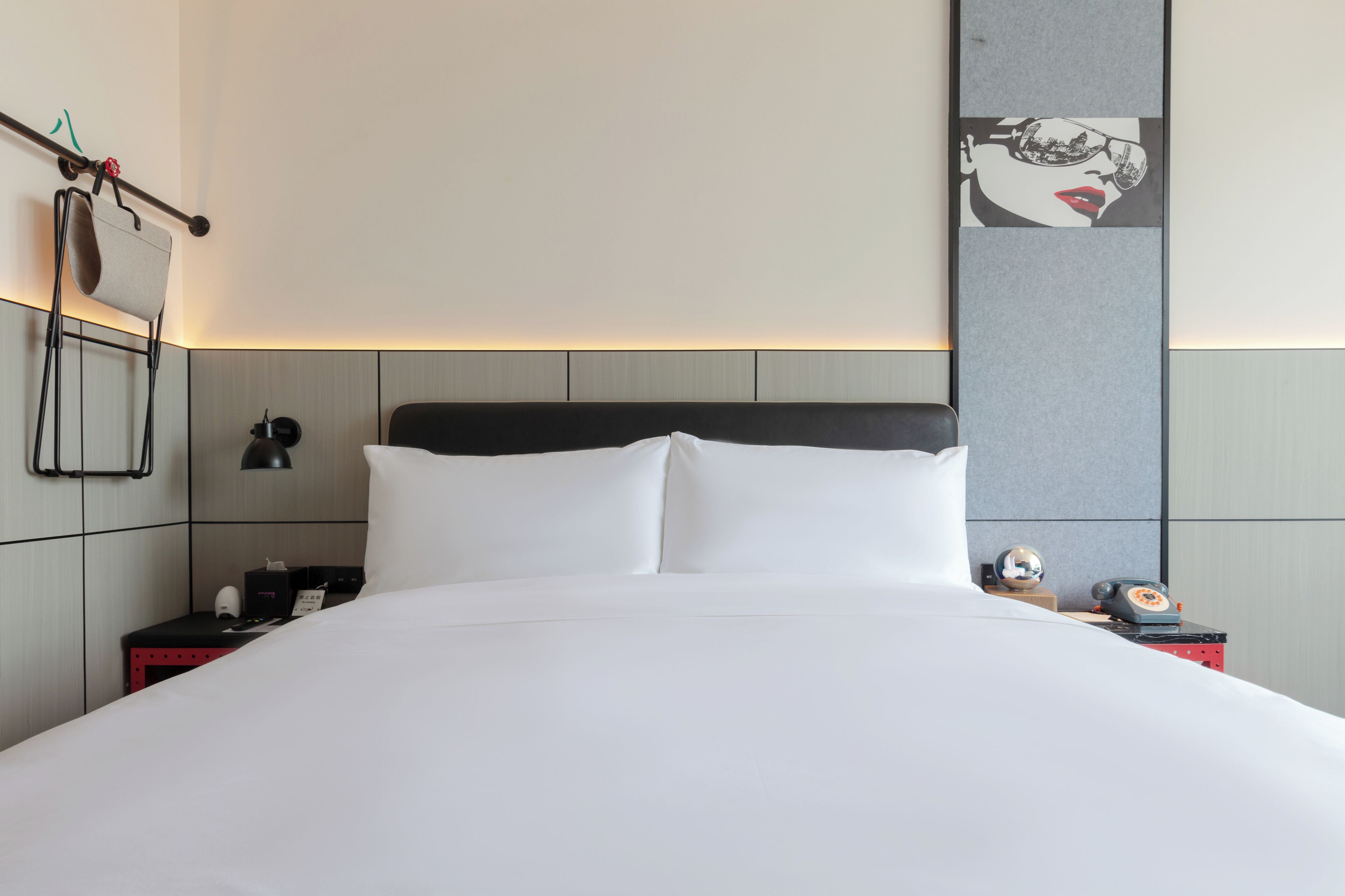 A hotel room with a large bed, a nightstand with a lamp, a small table with a globe and a telephone, and a wall-mounted towel rack.