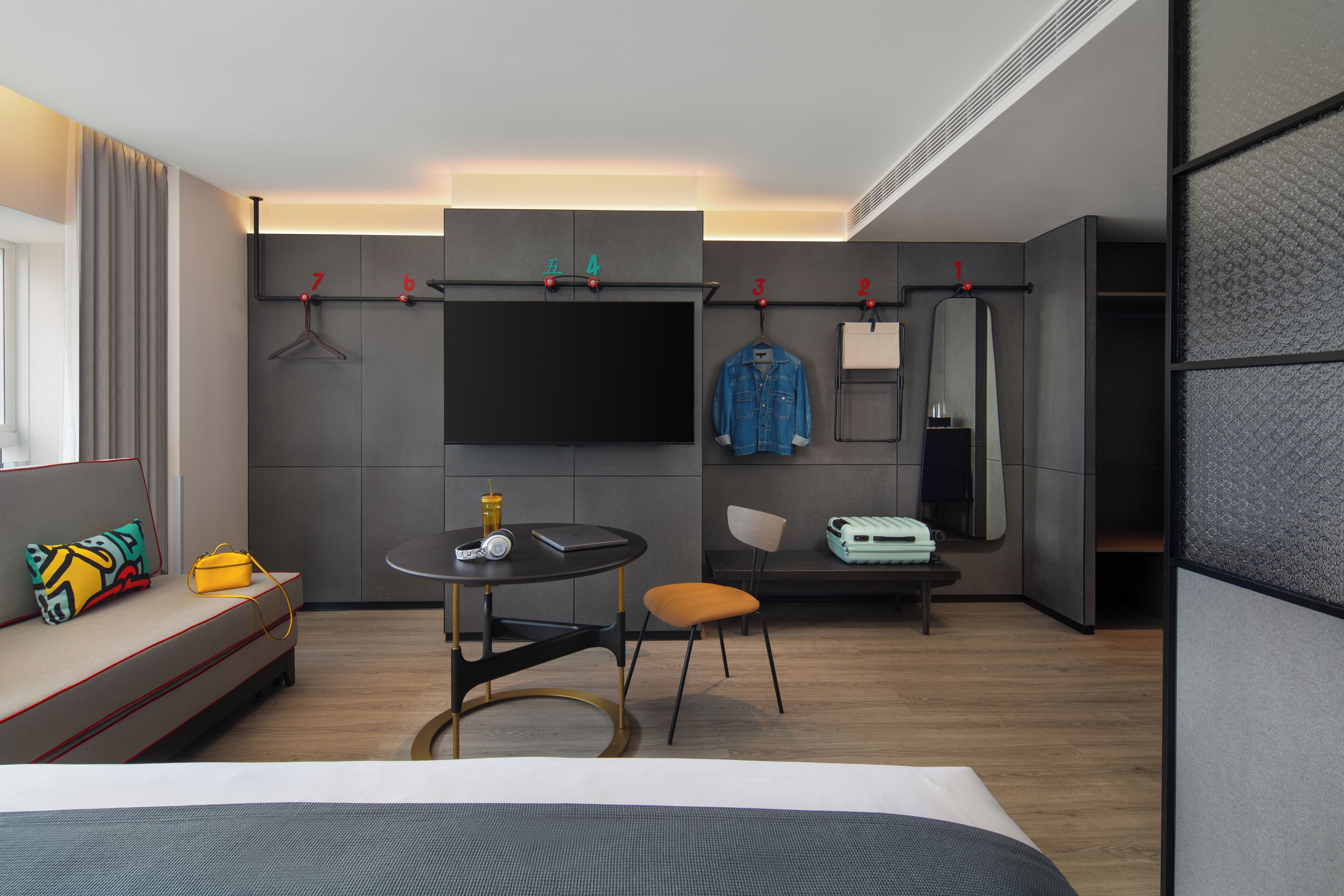 Quarto de hotel moderno com cama, escrivaninha, cadeira, televisão e cabideiro com cabides.