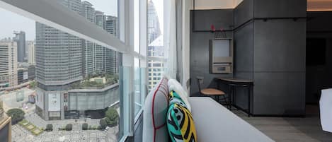 A modern hotel room with a large window overlooking a cityscape, a sofa with patterned cushions, and a minimalist kitchenette.
