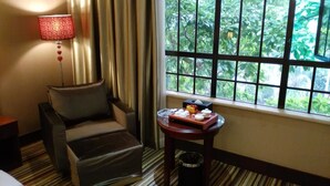 A hotel room with a leather armchair, a small table with a tray of items, a trash bin, and a window with a view of greenery. - Zhuhai Harmony Resort Hotel (Zhuhai)
