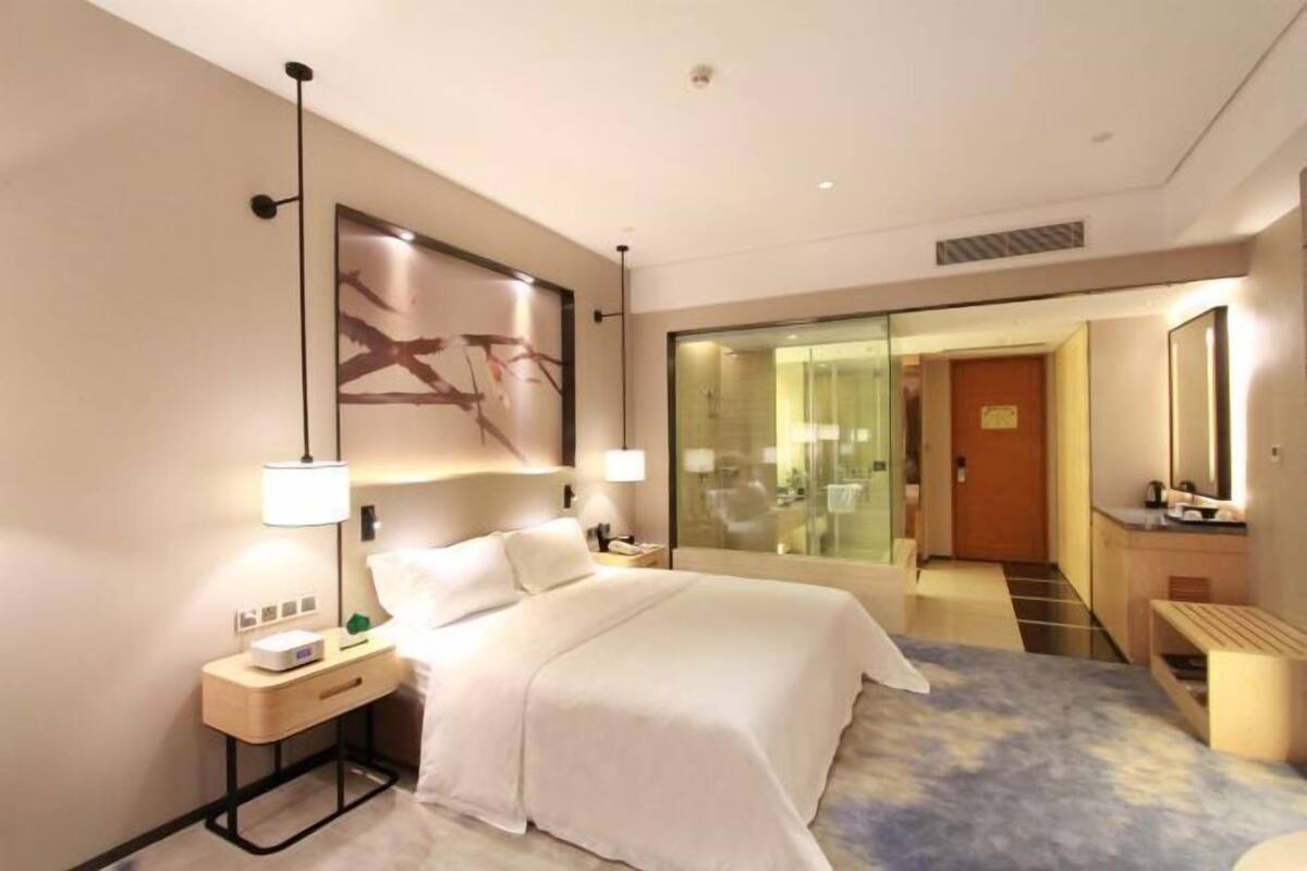 A modern hotel room with a large bed, bedside tables, a desk, and a glass partition.