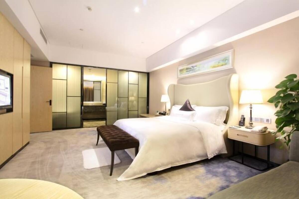 A hotel room with a bed, bedside tables, a bench, a TV, and a large mirror.