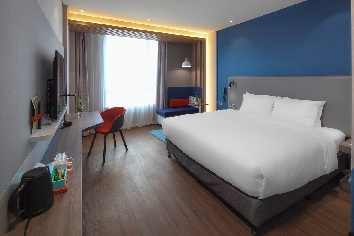 A hotel room with a large bed, a desk with a chair, a television, and a window with curtains.