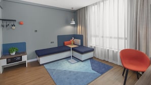 Une chambre moderne avec un canapé-lit, une petite table agrémentée d’une plante, un fauteuil rouge et une grande fenêtre ornée de rideaux légers.