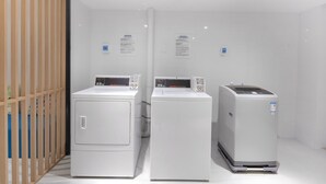 Une buanderie équipée de trois machines à laver et de trois sèche-linge industriels blancs, d’une cloison en lattes de bois et de murs recouverts de carreaux blancs.