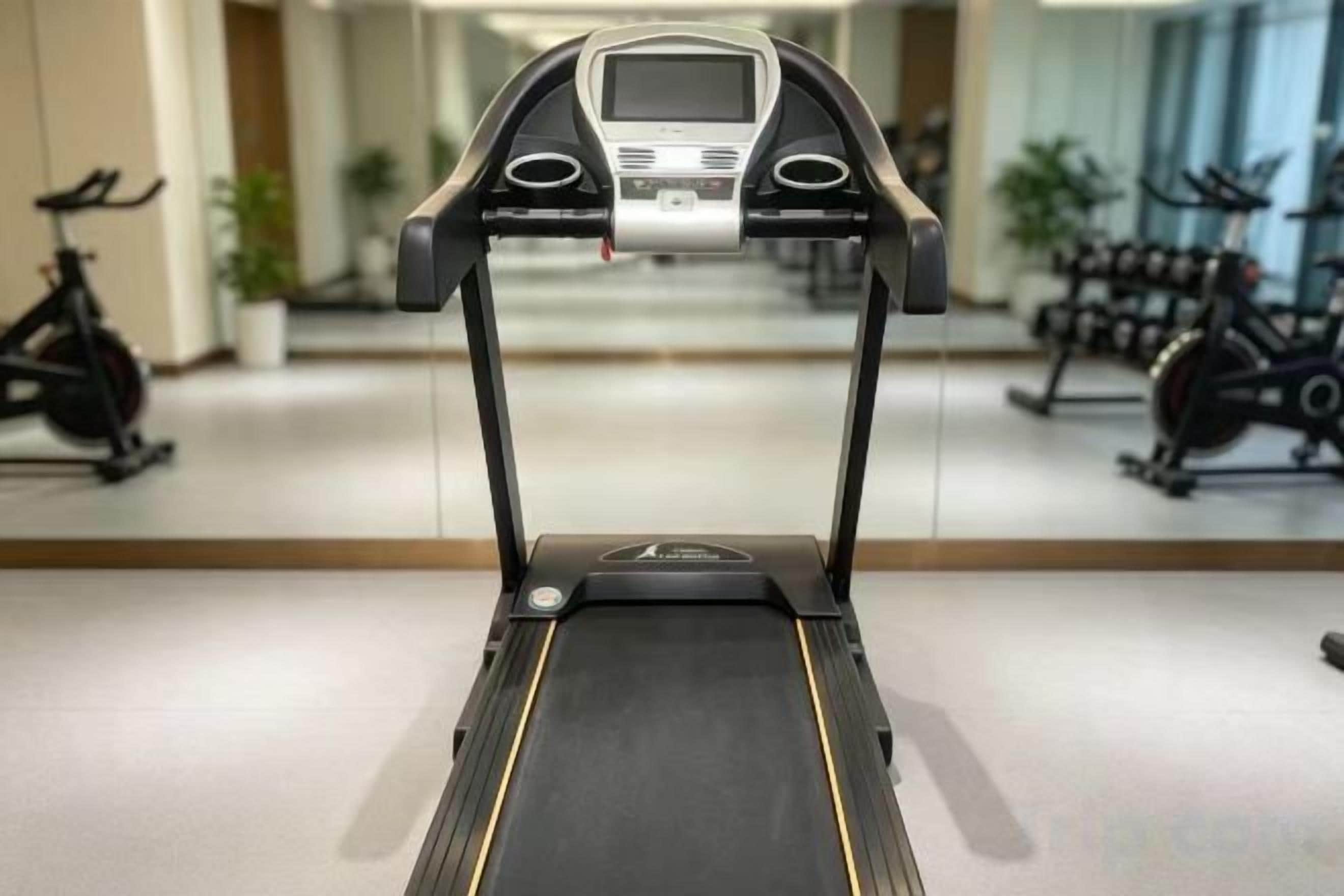 A treadmill in a gym with a mirrored wall and exercise bikes in the background.