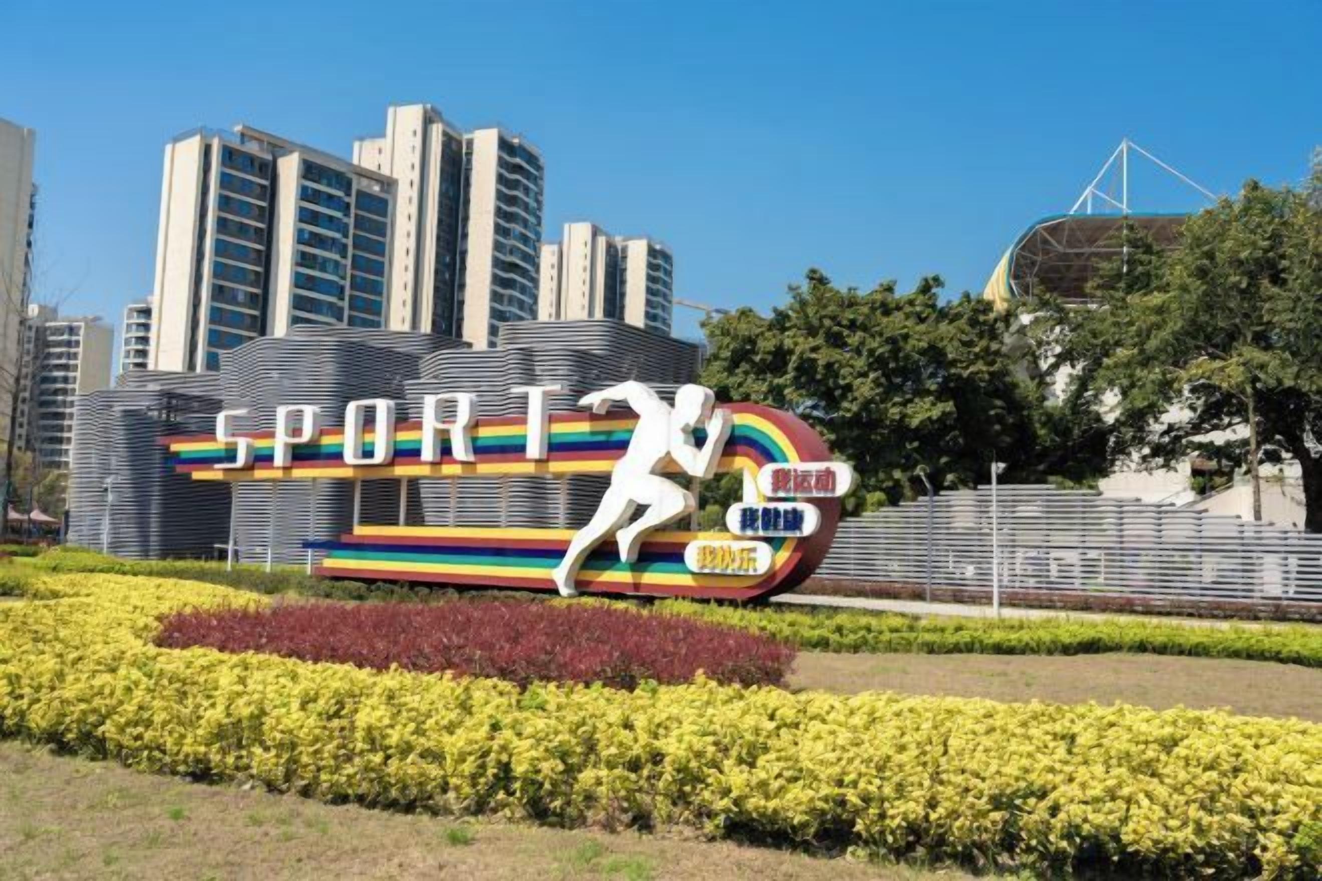 A sports-themed sculpture with the word 'SPORT' and a running figure, set against a backdrop of modern buildings and a clear sky.