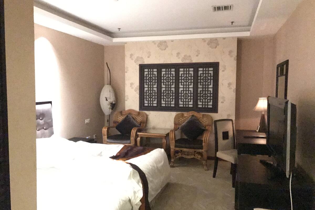 Habitación de hotel con una cama grande, dos sillones, un televisor y un biombo decorativo en la pared.