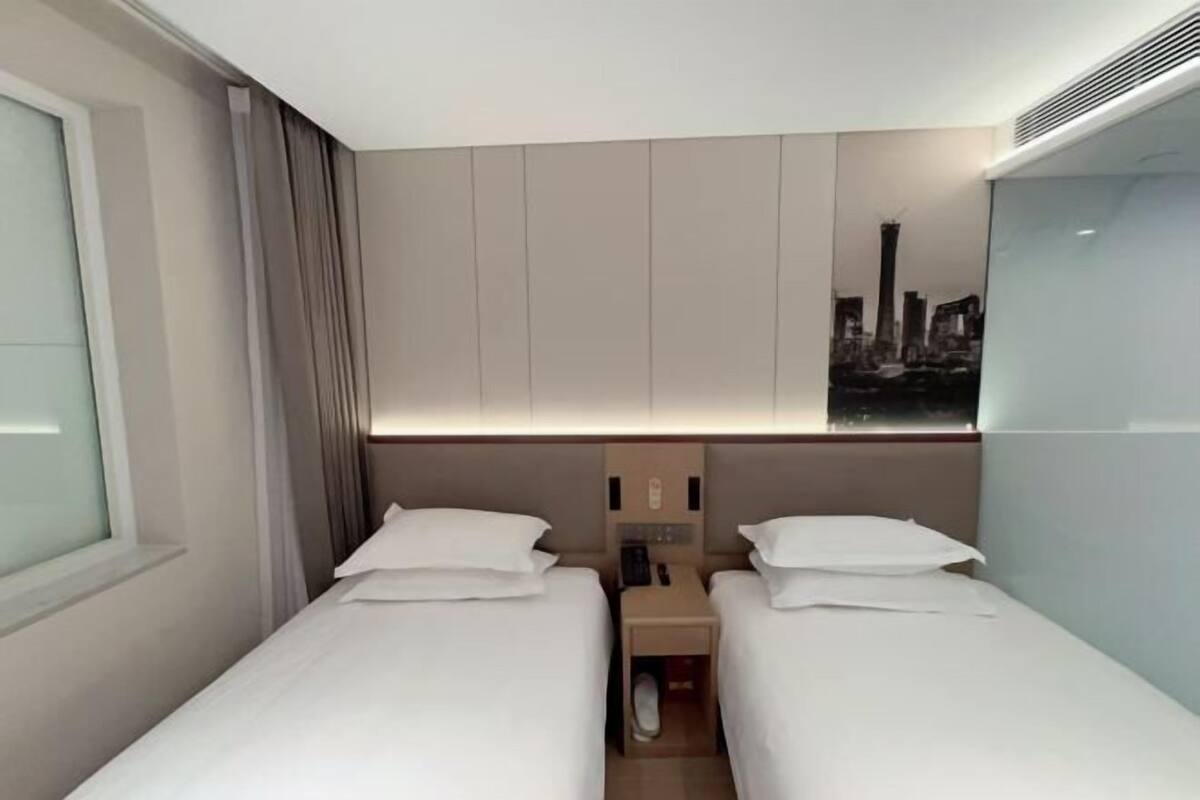 A hotel room with two beds, a nightstand, a window with curtains, a wall-mounted artwork, and a cityscape view.