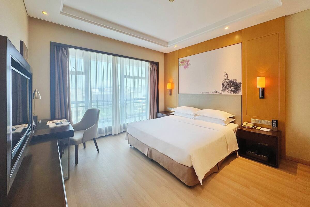 Habitación de hotel con cama, escritorio, silla y un ventanal con cortinas.
