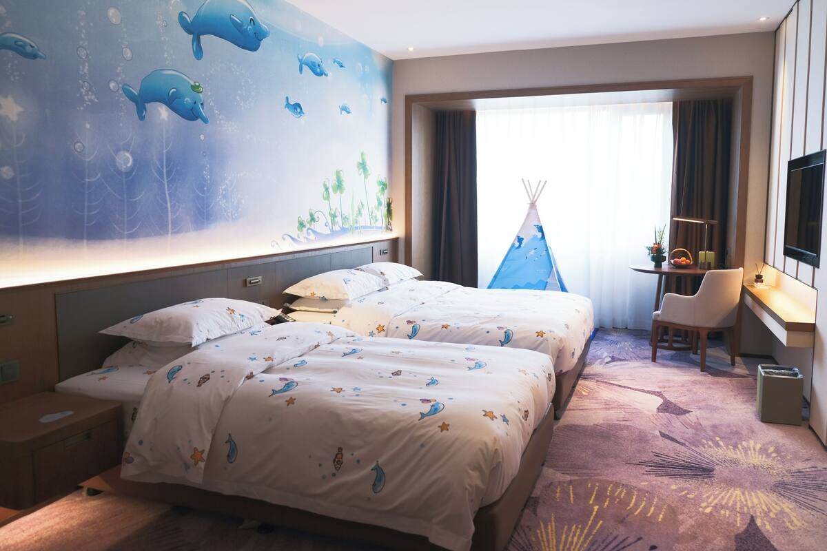 Habitación de hotel con dos camas, un escritorio, una silla, un televisor y un mural con criaturas marinas.