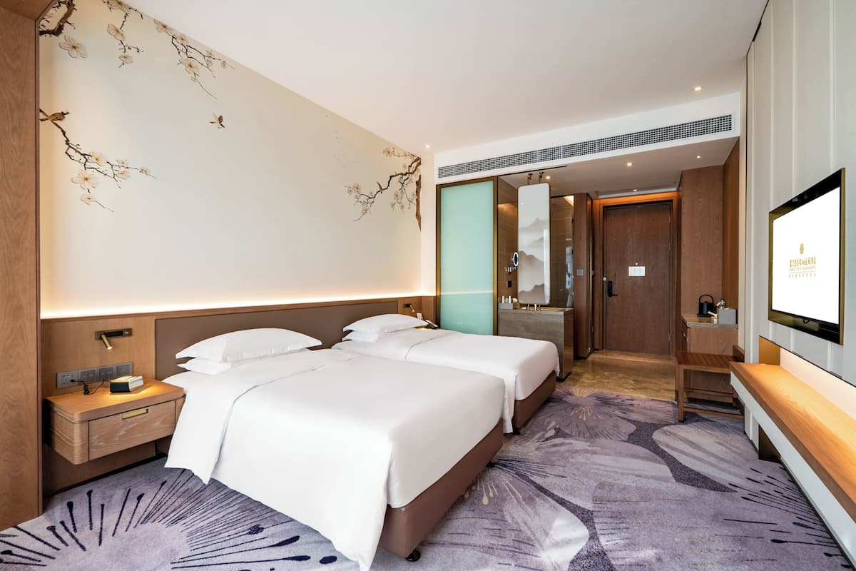 Habitación de hotel con dos camas, televisión, mesita de noche, espejo y baño visible a través de una puerta abierta.