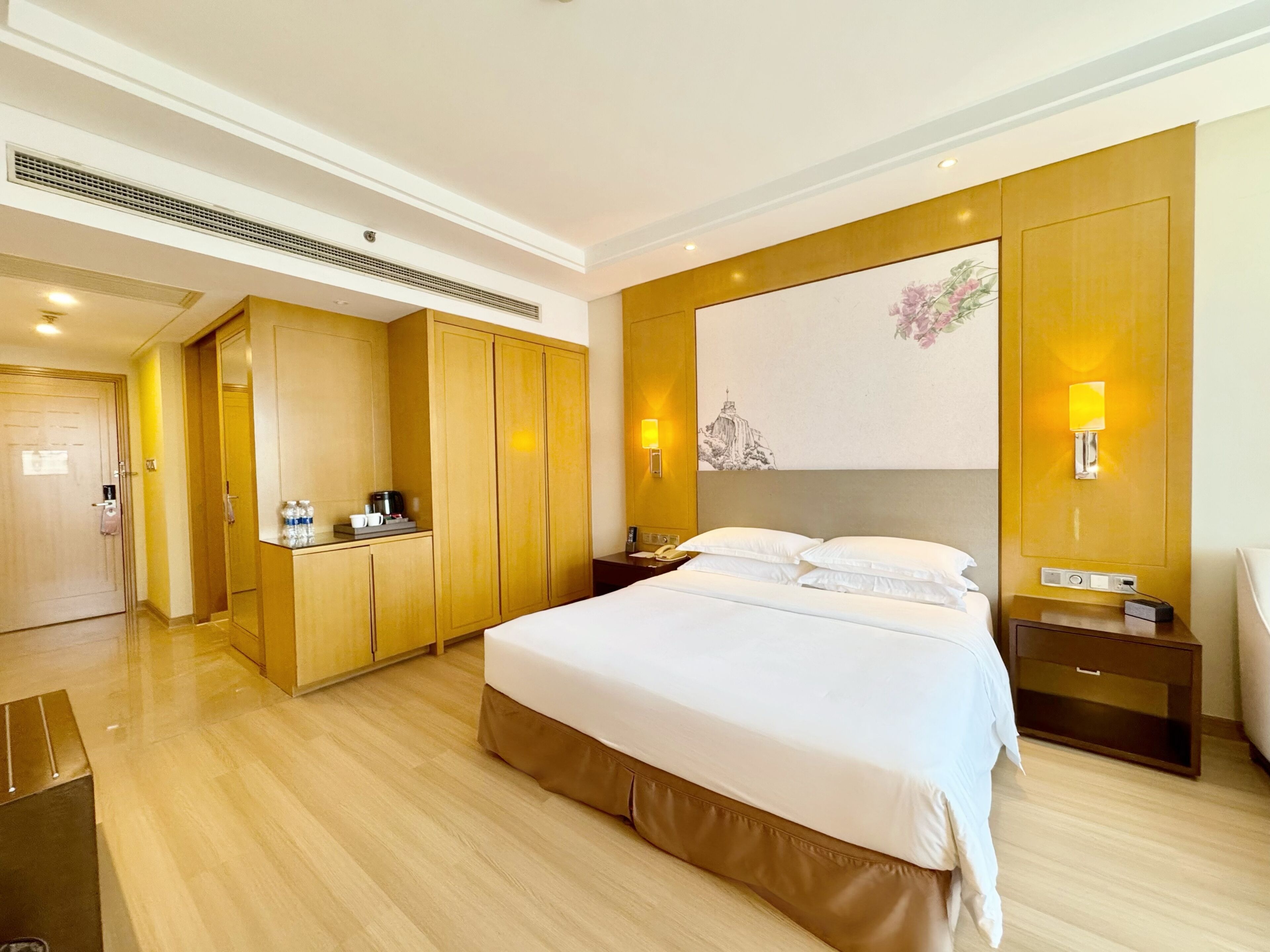 A hotel room with a large bed, wooden wardrobe, bedside tables, a nightstand, and a wall-mounted lamp.