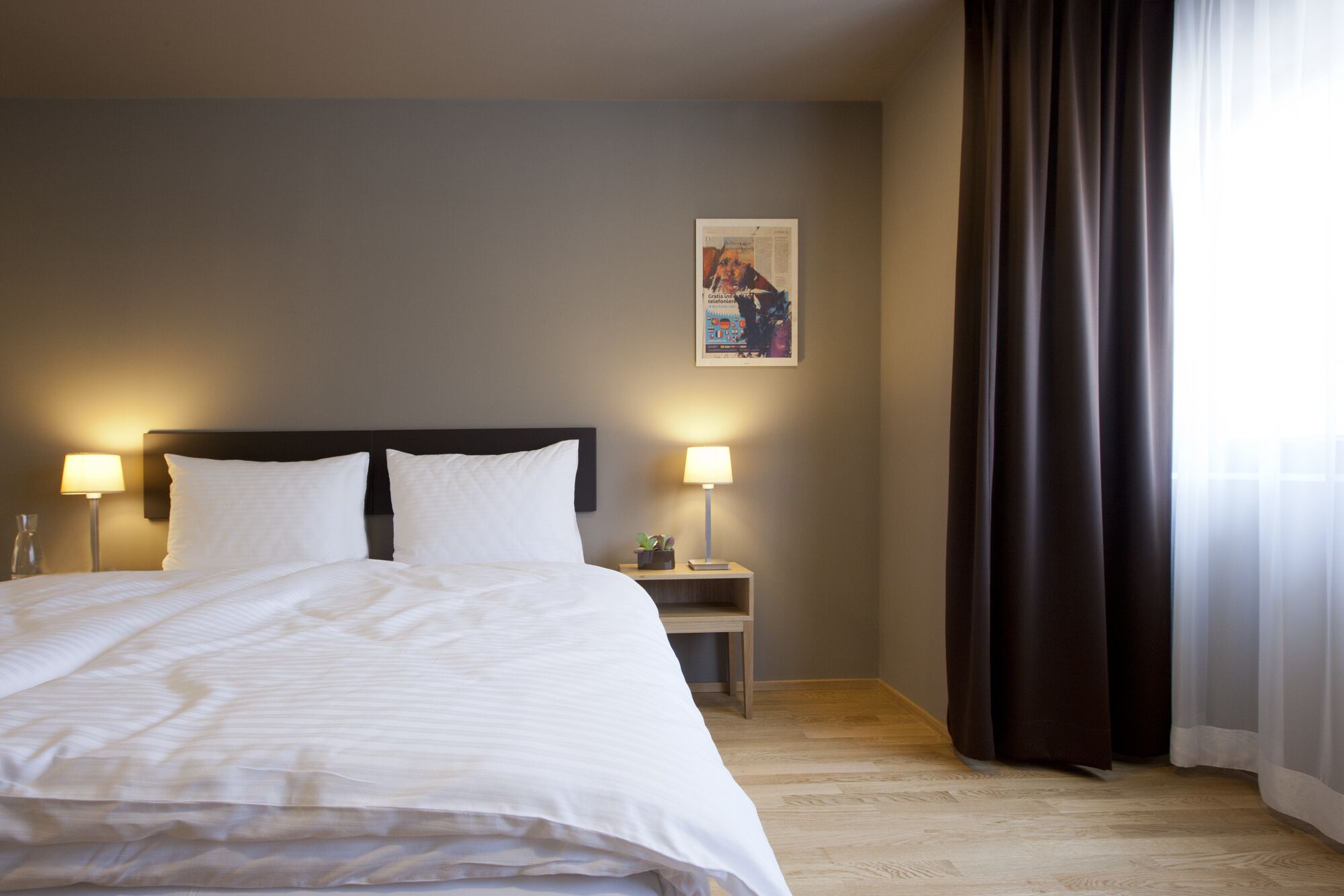 A hotel room with a bed, two bedside lamps, a nightstand, a framed picture, and a window with curtains.