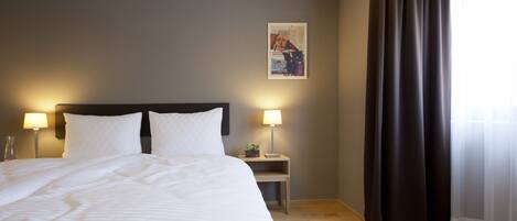 A hotel room with a bed, two bedside lamps, a nightstand, a framed picture, and a window with curtains.