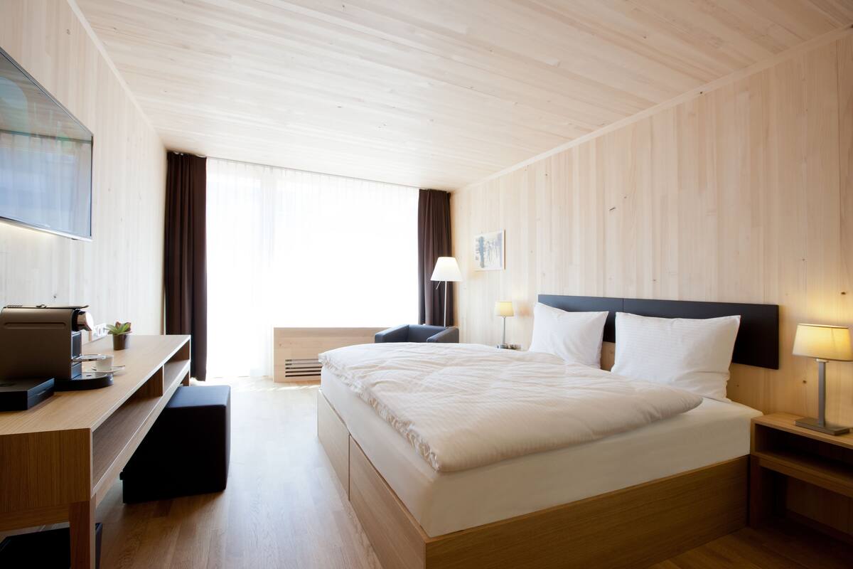 A hotel room with a bed, a desk with a computer, a chair, a nightstand with a lamp, and a window with curtains.