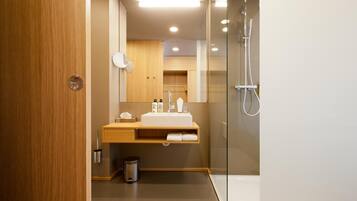 Un baño moderno con tocador de madera, cabina de ducha de vidrio y un cubo de basura visible.