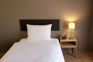 Una cama bien hecha con ropa blanca, un cabecero negro y una mesita de noche con una lámpara y una pequeña planta.