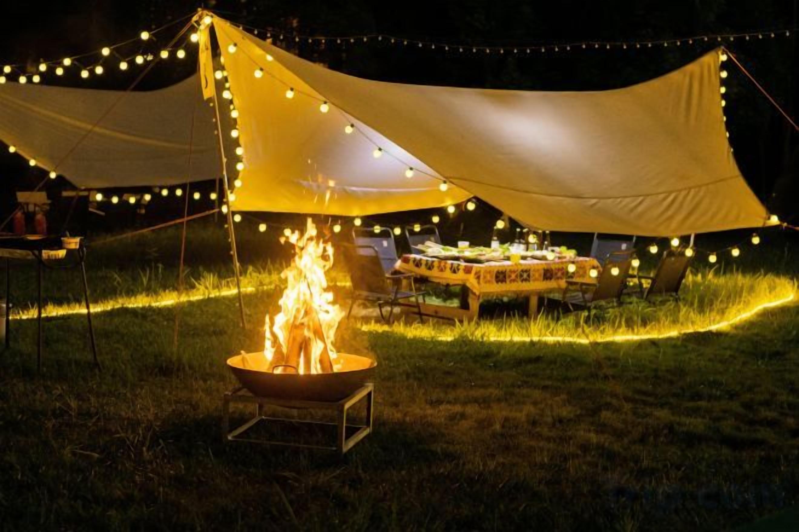 A tent with a fire pit and string lights, surrounded by a grassy area.