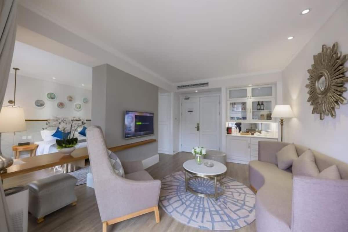 A modern living room with a sofa, armchair, and round coffee table. There is a TV mounted on the wall, a dining area with a table and chairs, and a kitchen area with cabinets and a countertop.