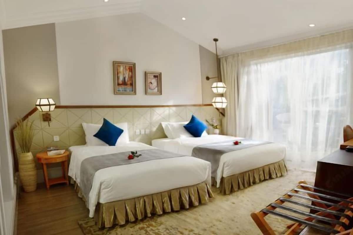 A hotel room with two beds, a small table, a chair, and a large window with sheer curtains.